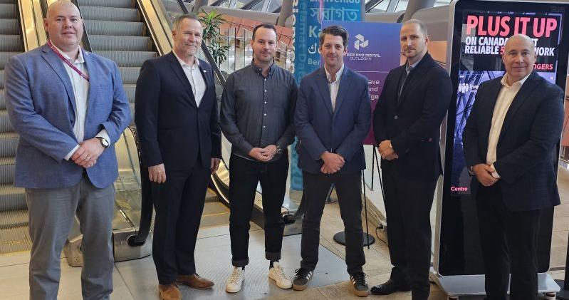 A group of six men stand side-by-side in an airport or mall-like setting near an escalator and digital signage. They are dressed in business or business-casual attire and facing the camera for a posed group photo