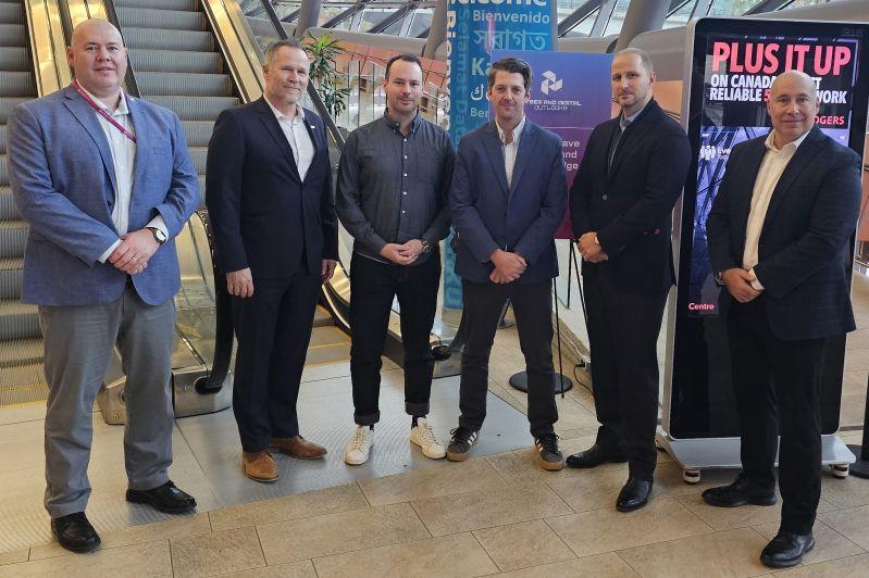 A group of six men stand side-by-side in an airport or mall-like setting near an escalator and digital signage. They are dressed in business or business-casual attire and facing the camera for a posed group photo