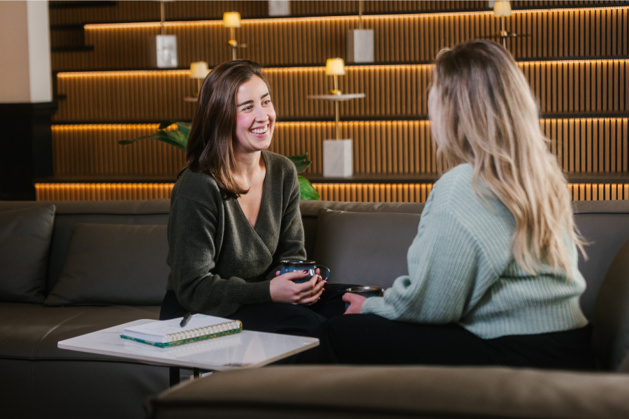 Two women sit on a couch in a modern lounge, smiling and talking while holding coffee mugs, with a notebook on the table between them.