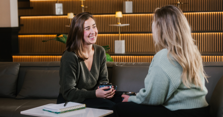 Two women sit on a couch in a modern lounge, smiling and talking while holding coffee mugs, with a notebook on the table between them.