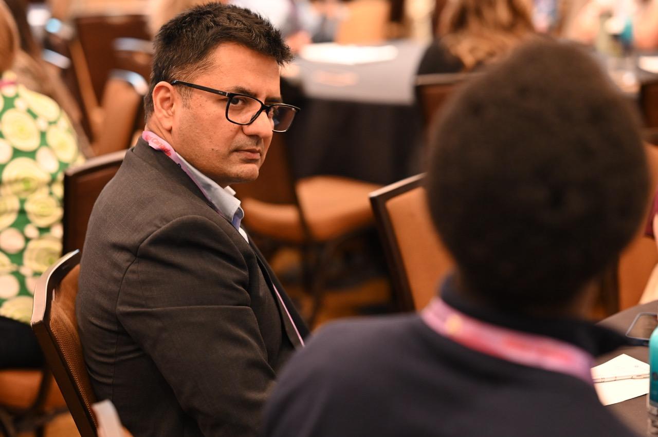 A man with dark hair and glasses, wearing a suit jacket and a light blue collared shirt, is looking over his left shoulder directly at the camera. He has a serious expression on his face. A lanyard is visible around his neck. In the foreground, the back of another person's head and shoulders are visible, out of focus. The background shows a blurred conference or event setting with other people and tables.