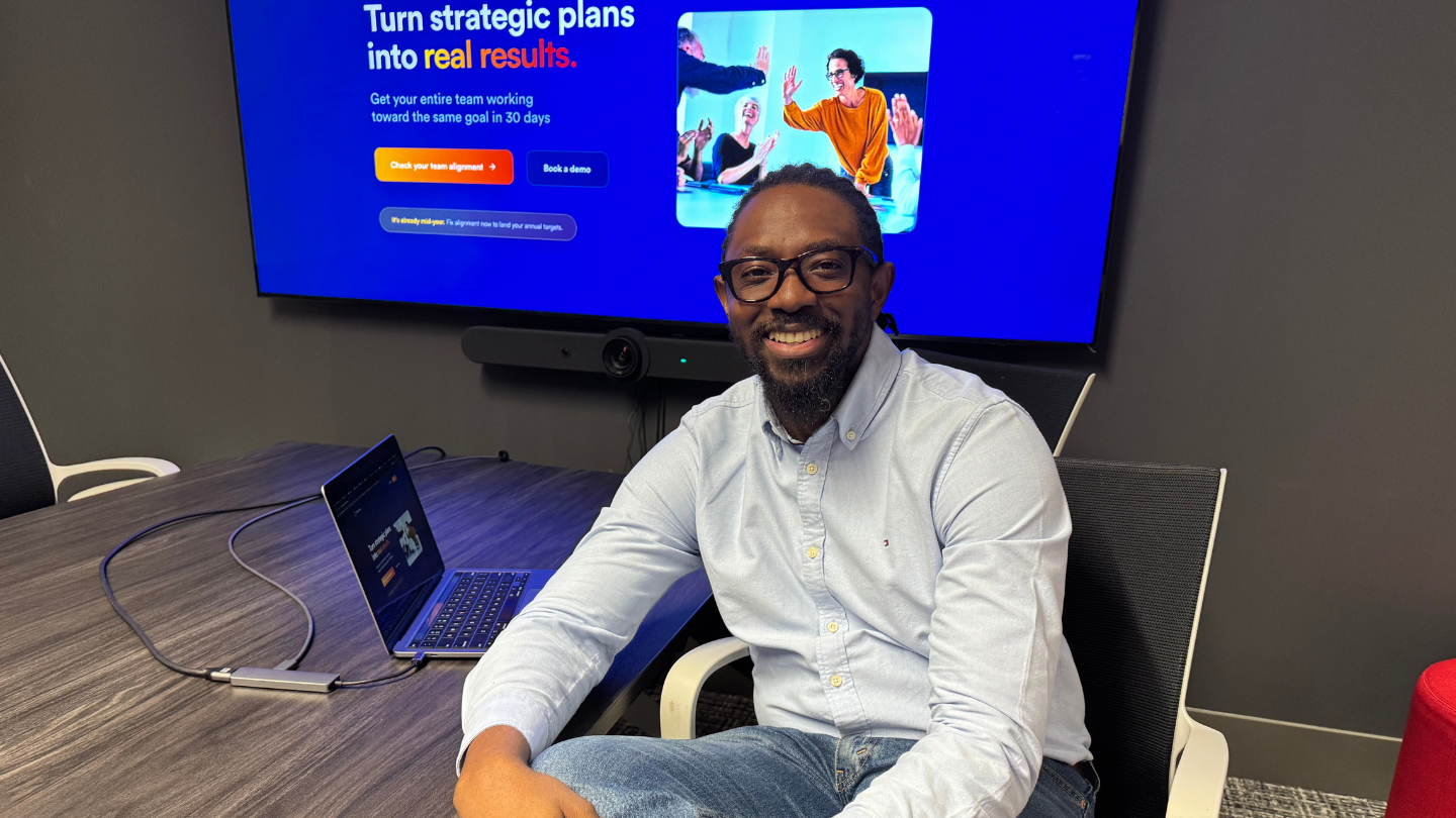 Tom Olajide, founder of MissionSync, sits smiling at a conference table with his laptop open. Behind him, a large screen displays the MissionSync website with the words “Turn strategic plans into real results.”