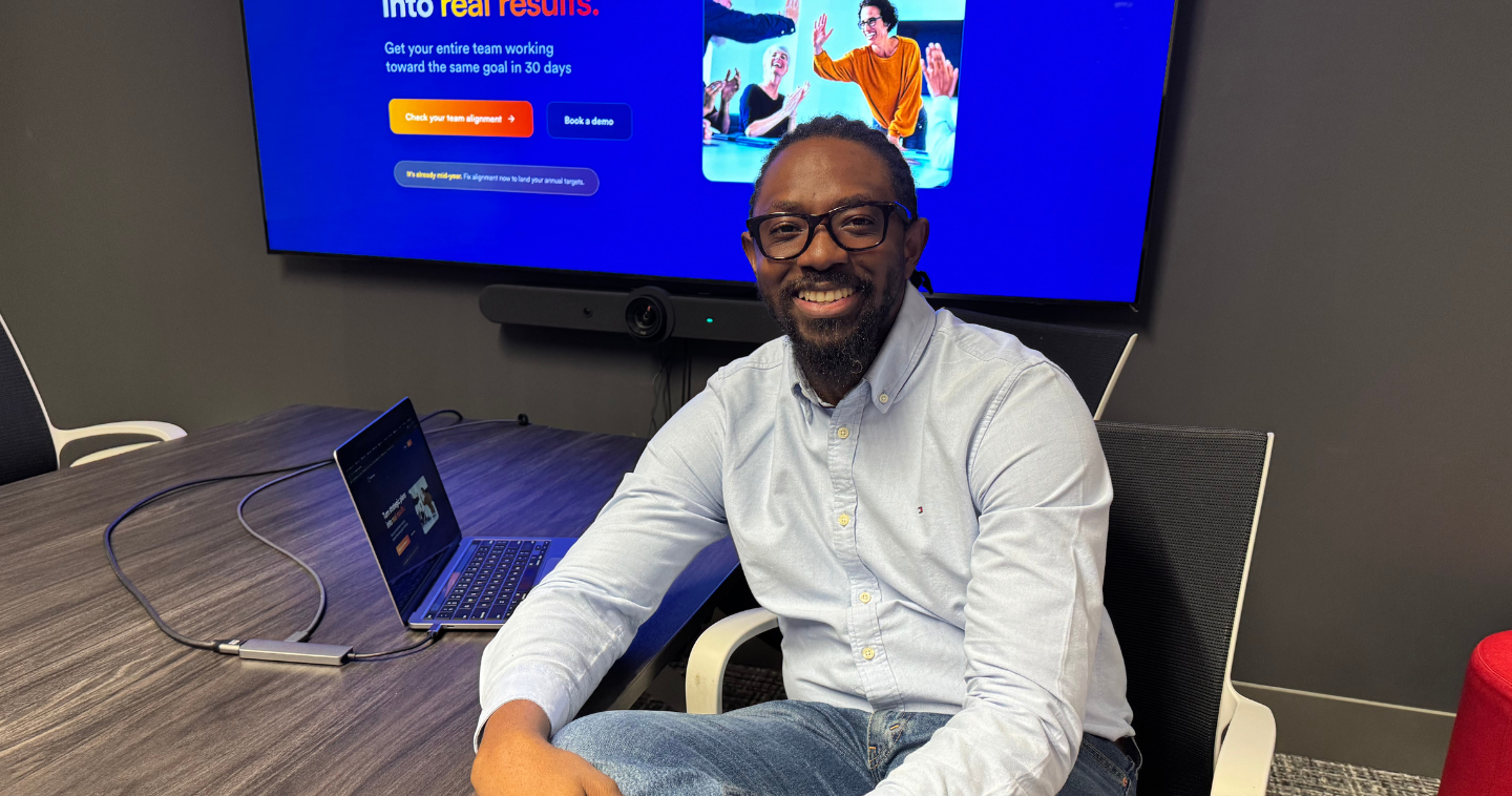 Tom Olajide, founder of MissionSync, sits smiling at a conference table with his laptop open. Behind him, a large screen displays the MissionSync website with the words “Turn strategic plans into real results.”