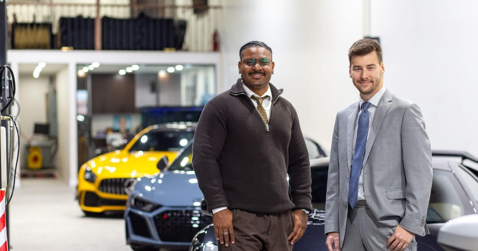 Two people stand smiling in a luxury car showroom. One person wears glasses, a brown sweater, and brown trousers; the other wears a light grey suit with a blue tie. Behind them are several high-end sports cars, including a yellow car, a grey car, and a dark-colored Lamborghini. The setting appears bright and modern, with office space visible in the background.