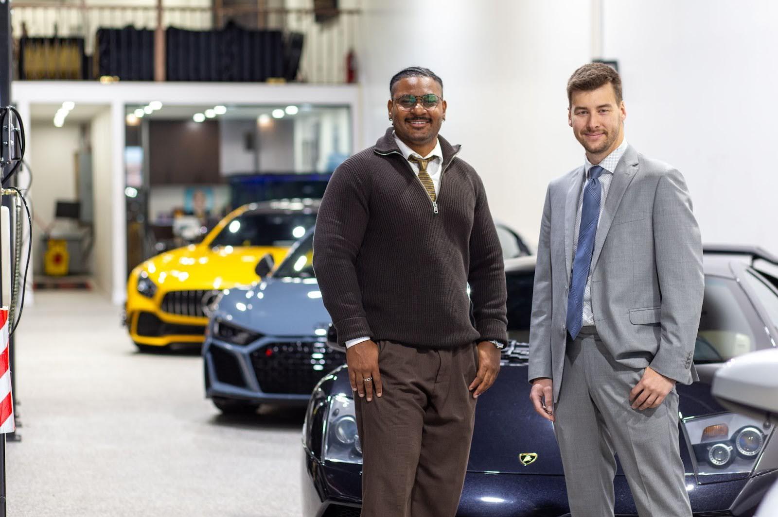 Two people stand smiling in a luxury car showroom. One person wears glasses, a brown sweater, and brown trousers; the other wears a light grey suit with a blue tie. Behind them are several high-end sports cars, including a yellow car, a grey car, and a dark-colored Lamborghini. The setting appears bright and modern, with office space visible in the background.