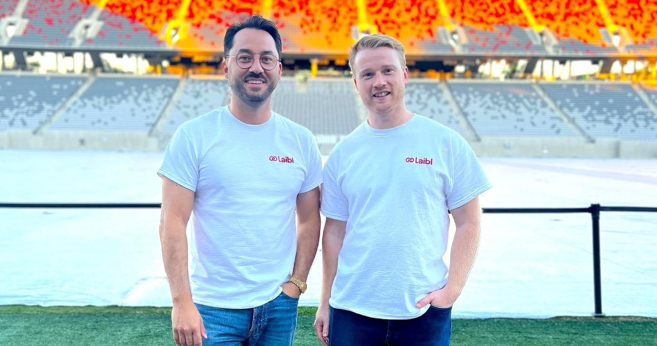 Two men standing on a green field in a large sports stadium with red and orange seating, both wearing white t-shirts with the Laibl logo.